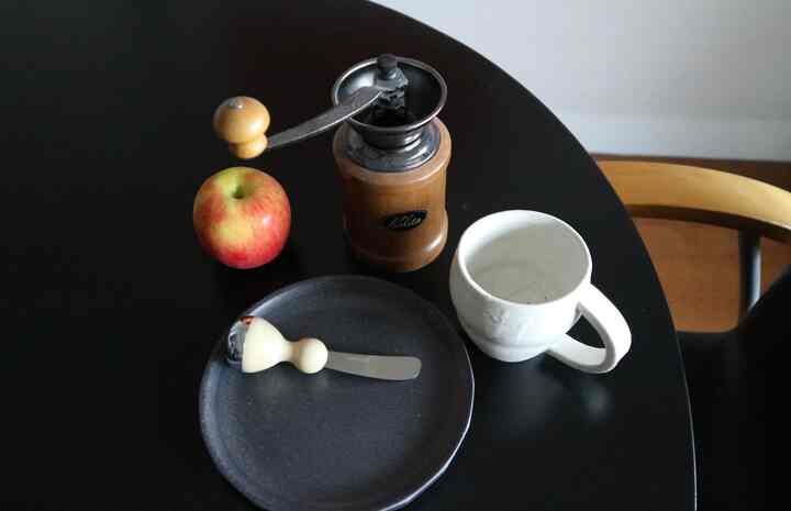 黒い丸テーブルの上にコーヒーミル、リンゴ、マグカップ、皿、バターナイフが置かれた朝食の雰囲気ある空間