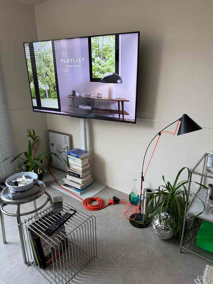Simple living room with light gray flooring and white walls featuring blinds, TV stand, floor lamp, and multiple plants
