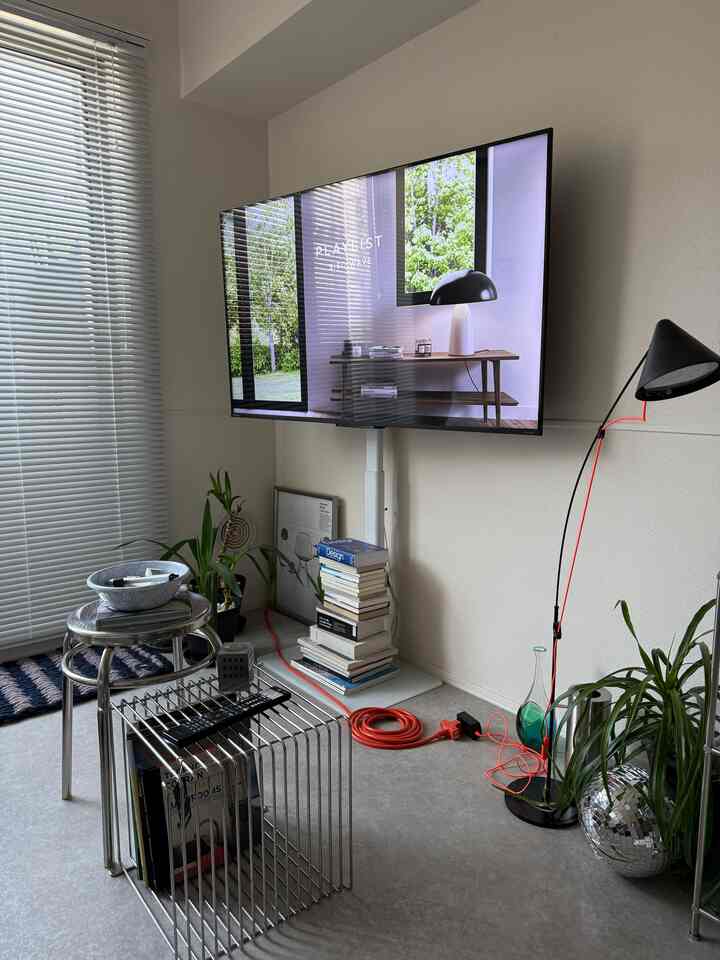 Living room in white and gray tones featuring a wall-mounted TV, metal shelves, blinds, floor lamp, and plants in a minimalistic setting