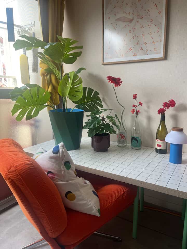 グリーンの植物と赤い花が配置されたミッドセンチュリーモダンの1LDK空間、オレンジのアームチェアと白い格子柄のテーブルが居心地の良い雰囲気を作り出すインテリア