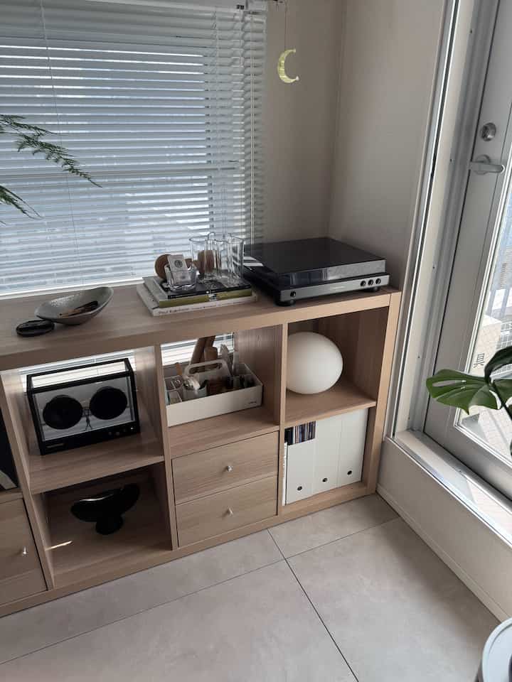 明るい木目調の棚とブラインドのある窓辺に、レコードプレーヤーと透明スピーカーが配置された北欧風のリビングスペース