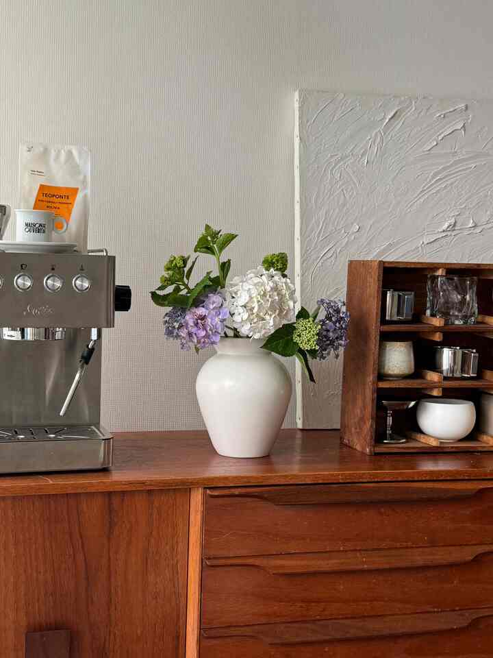 Natural tone space featuring a brown wooden sideboard topped with a white vase of hydrangeas and a coffee machine against white walls