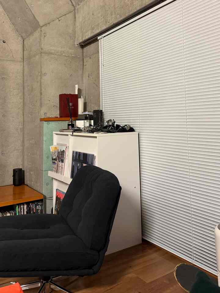 グレーの露出コンクリート壁と白いブラインドがある空間に、黒いアームチェアと白い収納キャビネットが配置されたインダストリアルな雰囲気のインテリア