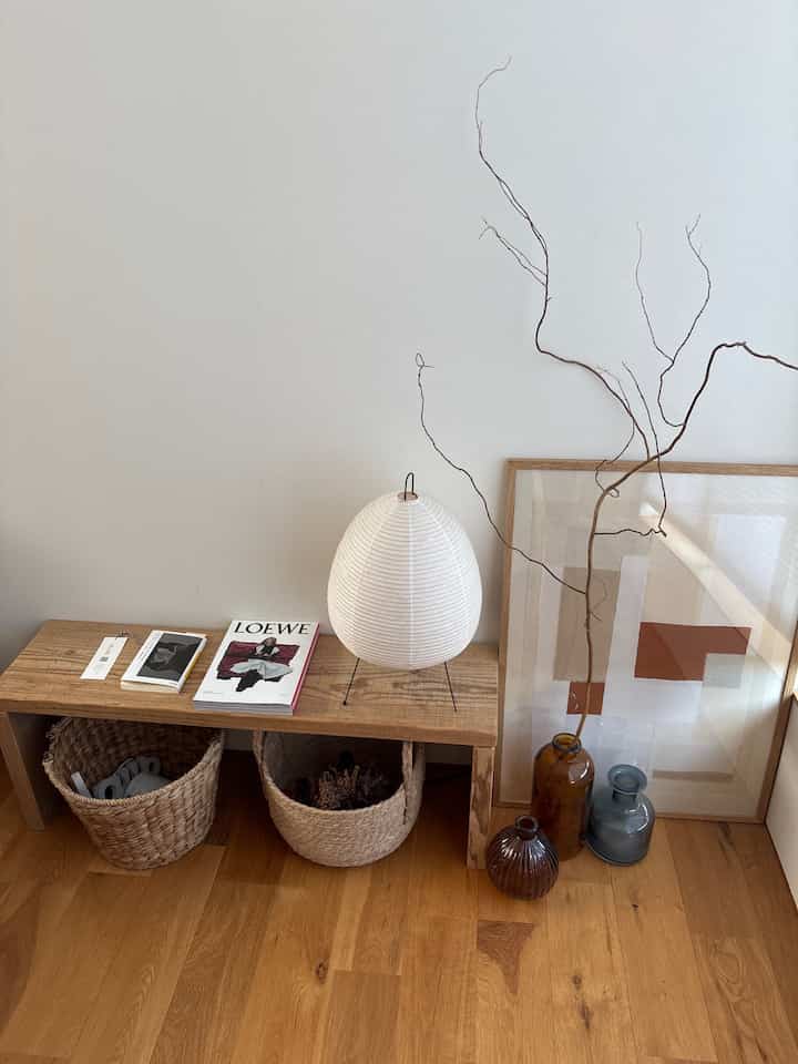 落ち着いたベージュとウッドトーンの寝室で、木製ベンチと2つのラタンバスケット、アカリのスタンドランプが配置された雰囲気のあるインテリア。