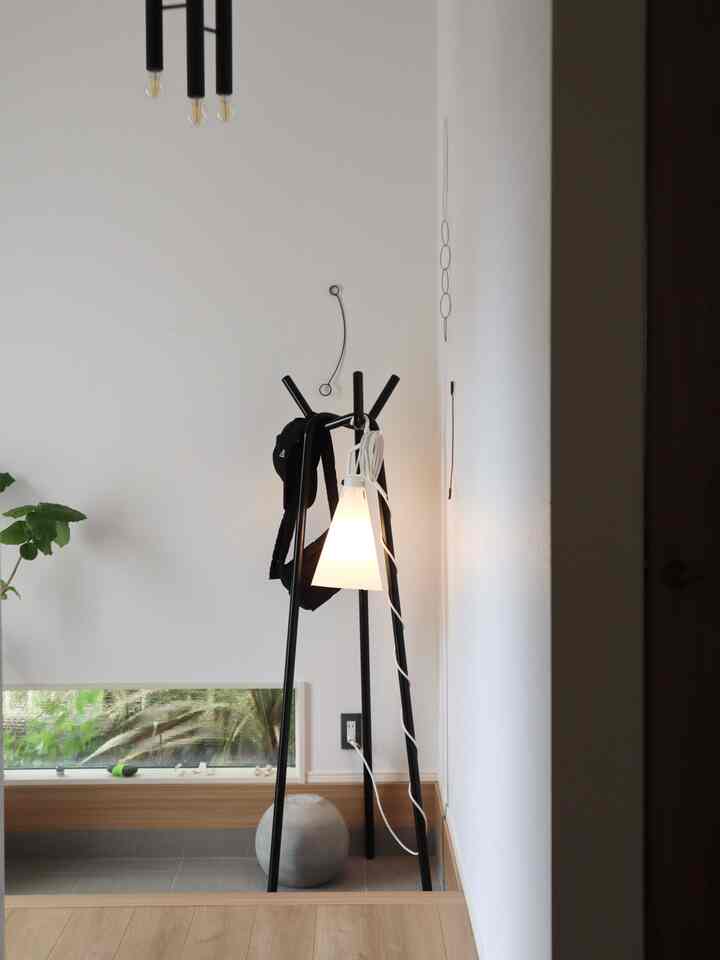 白い壁とナチュラルウッド調の床が調和する玄関、中央に黒いコートハンガーとランプが配されたミニマルなインテリア