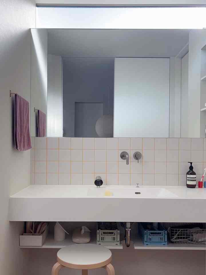 白とベージュのタイル調が調和するバスルームの洗面スペースで、シンプルで整頓された収納と木製のスツールが配された清潔感のあるインテリア