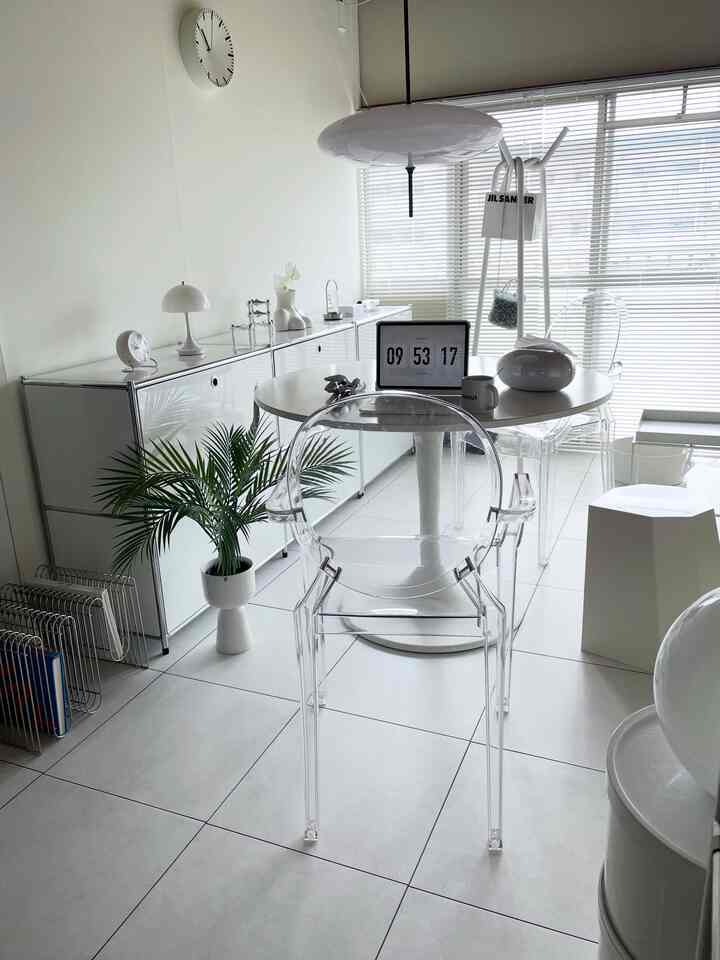 白を基調としたモダンなワンルームリビング、透明なルイゴーストのアームチェアと丸いダイニングテーブル、整然としたキャビネットとグリーンプラントが配置された雰囲気のあるインテリア