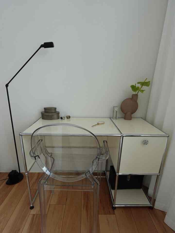 A white-toned wall and wood floor blend in a space featuring a transparent chair and steel frame desk, creating a sleek modern workspace