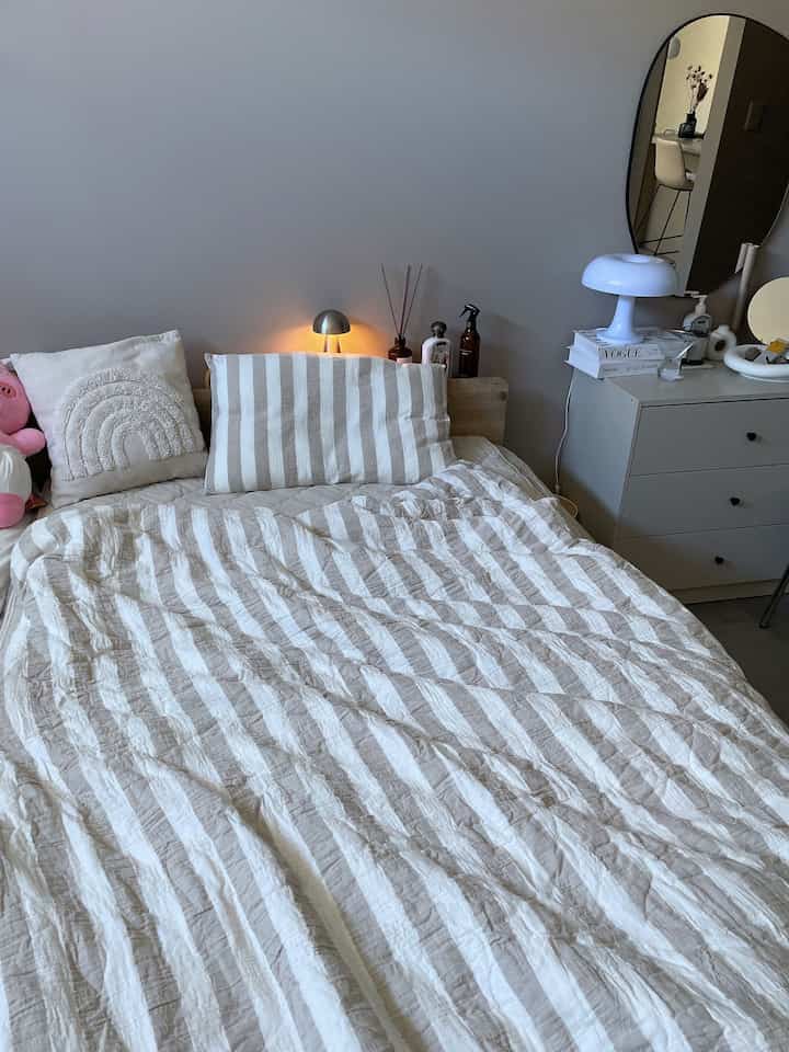 Beige and white toned bedroom featuring striped duvet cover, nightstand, and round mirror creating a cozy atmosphere
