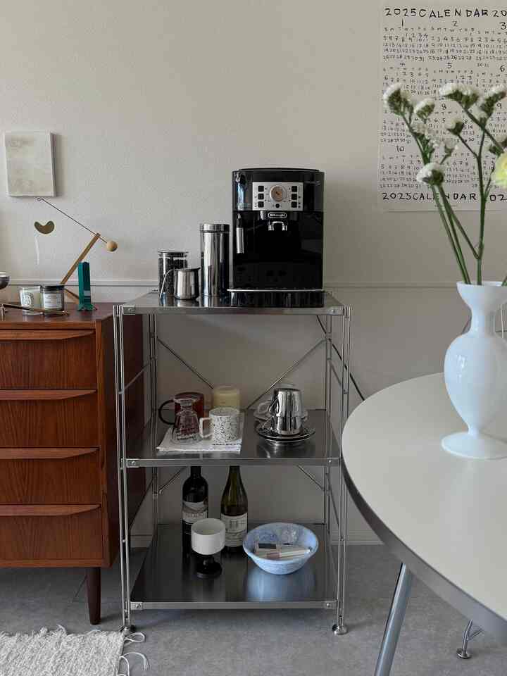 白い壁とシルバーのステンレスユニットシェルフがあるナチュラルモダンなダイニングルーム、中央に黒いエスプレッソマシンとコーヒーアクセサリーが配置された印象的な空間