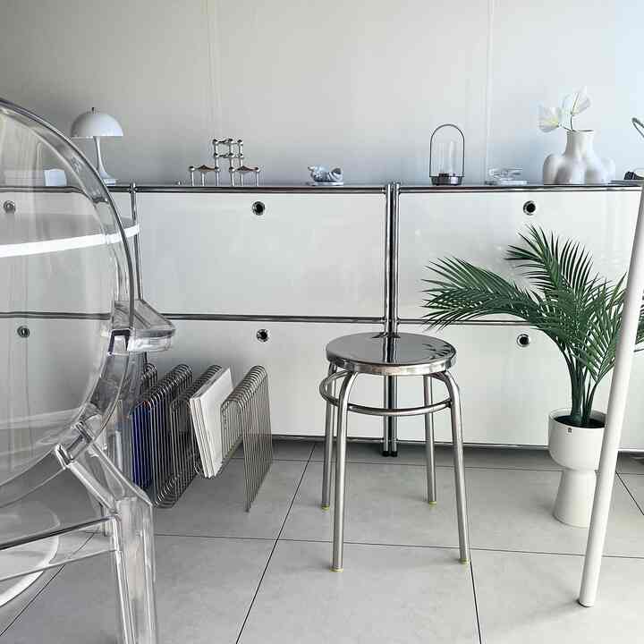 A compact room in white and silver tones featuring a stainless round stool at center and a plant, creating a minimal and natural interior