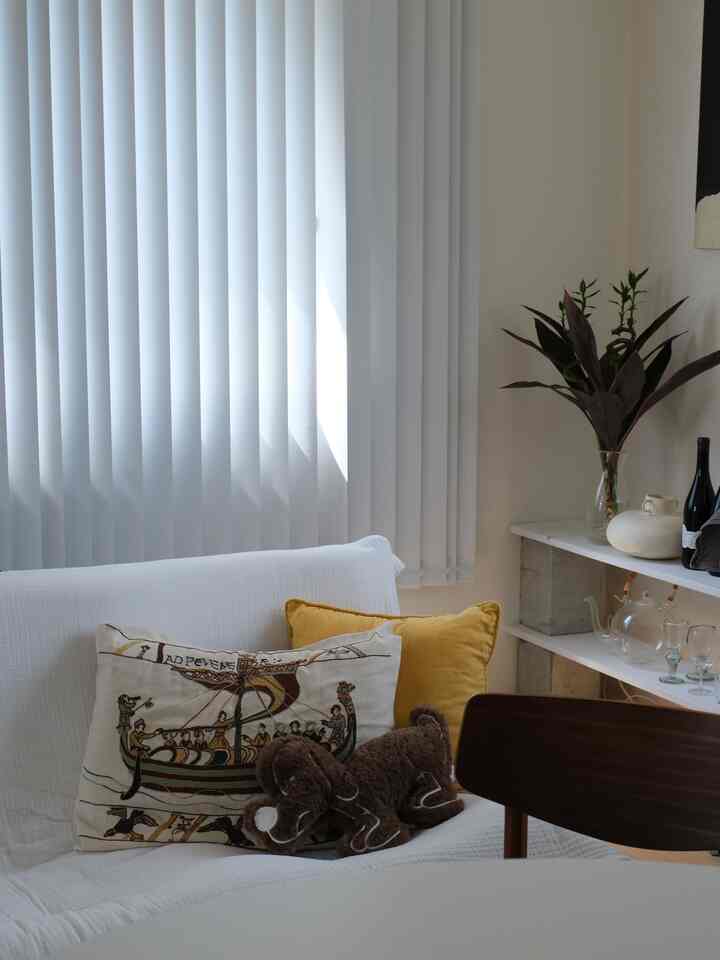 白と茶色のトーンで構成された1LDKのリビング、白いソファにヴィンテージ刺繍クッションが置かれ、バーチカルブラインドが配置された雰囲気のあるインテリア