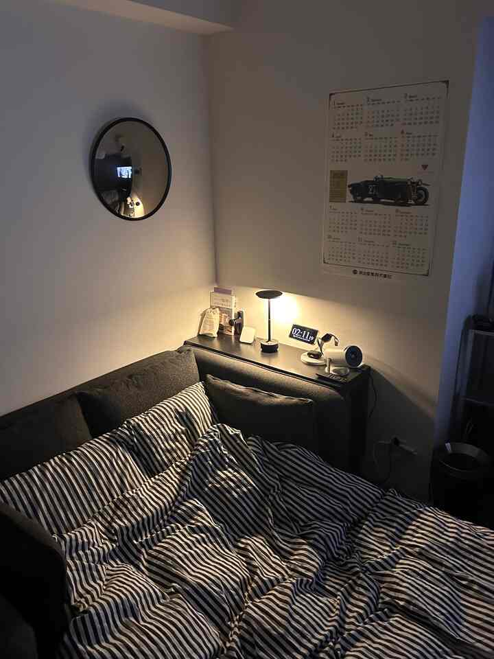 白い壁と黒いソファベッドがあるコンパクトなワンルーム寝室、間接照明と丸い壁鏡が居心地の良い雰囲気を演出