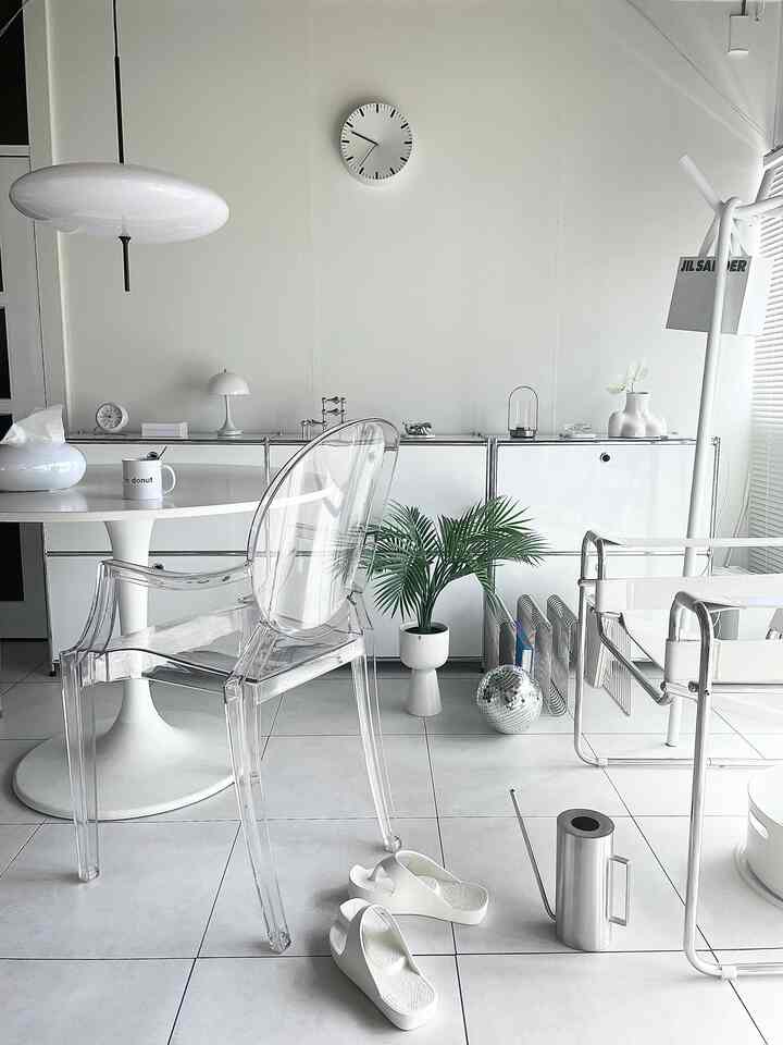 白とシルバーのトーンのリビング・ダイニングルーム、透明なアームチェアとステンレス製のじょうろが配置されたモダンで清潔感のあるインテリア