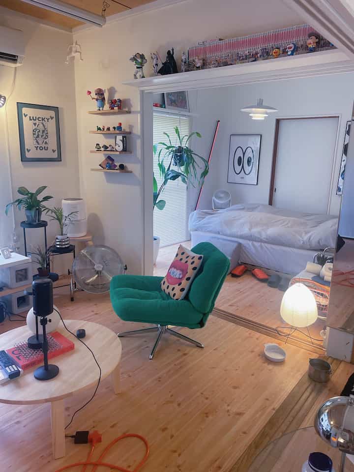 Natural color and white toned living room connected to bedroom, featuring a green armchair, coffee table, and potted plants in a cozy atmosphere