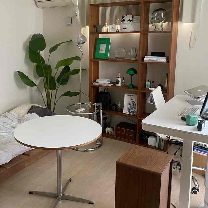 白と木目調が調和したワンルーム寝室、カフェ風のテーブルと本棚、フェイクグリーンが映えるシンプルモダンなホームオフィス