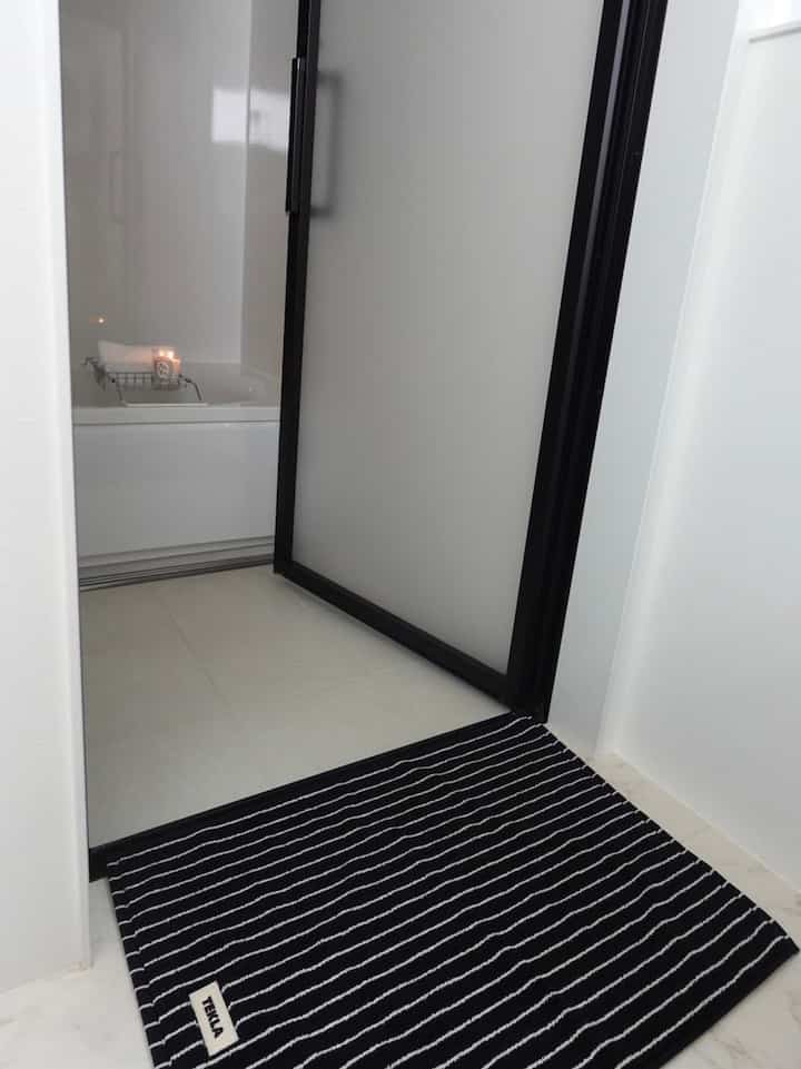 Minimalist bathroom in white and black tones featuring a bathtub with candle on tray and a striped bath mat, creating a clean atmosphere