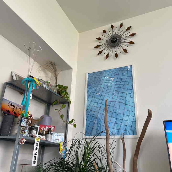 白い壁とミッドセンチュリーモダンの装飾時計があるリビング、大きな青いポスターと植物が配置された雰囲気のあるインテリア