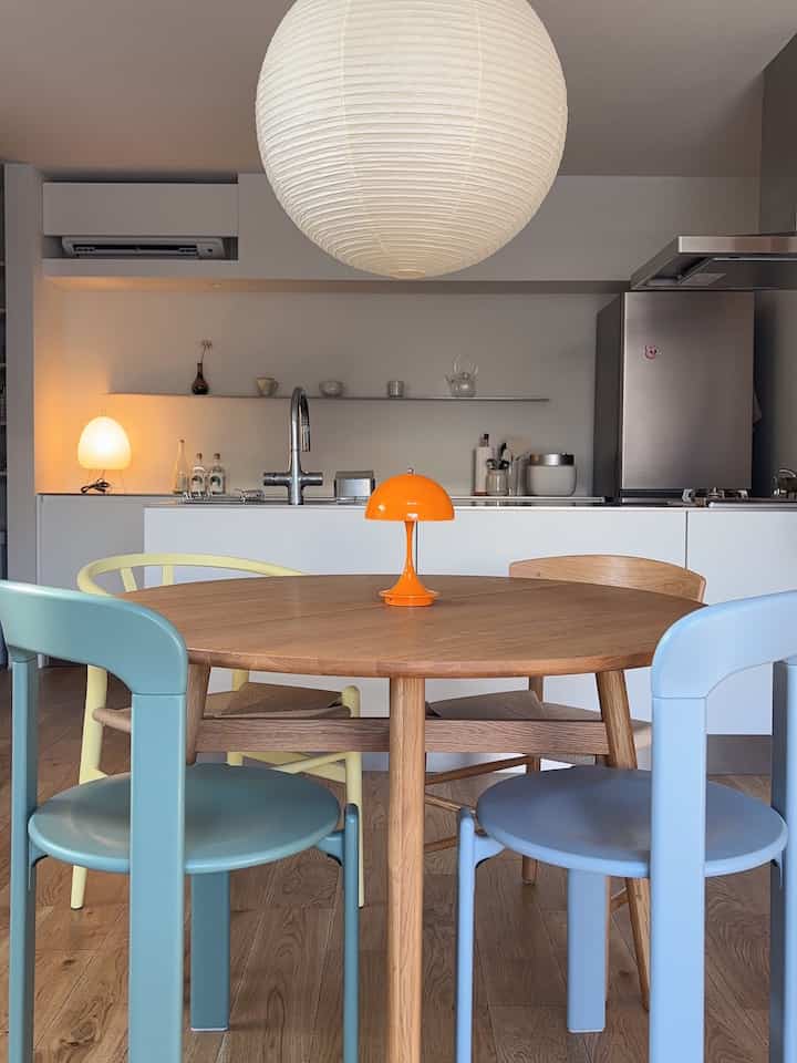ナチュラルな色調のダイニング、木製テーブルとパステルカラーの椅子、オレンジのテーブルランプが配置された印象的な空間