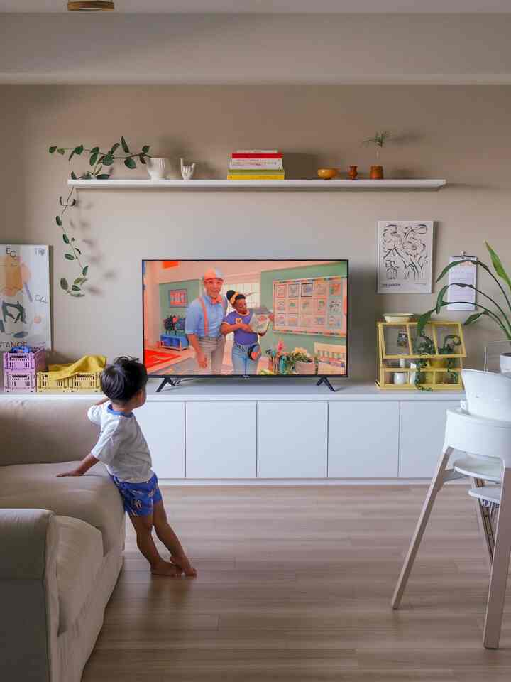 ナチュラルな木目調の床とホワイト家具が配置されたリビング、ソファ横に立つ子供がテレビを観る穏やかな空間