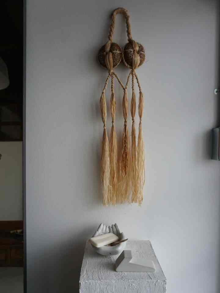 ナチュラル調の玄関空間、壁中央に自然素材の吊り飾りが配置され、下部には白いオブジェが置かれたミニマルで落ち着いたインテリア