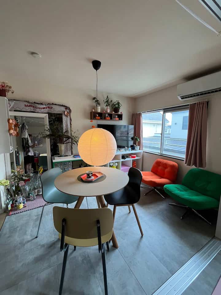 ベージュ調の壁とカーテンがある1LDKのリビング、円形のダイニングテーブルとカラフルな4脚の椅子、ペンダントライトが配された雰囲気のあるインテリア