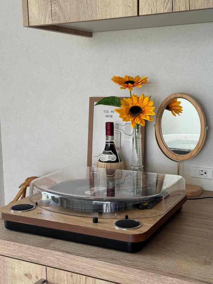 ベージュの壁と木目調のキャビネットにレコードプレーヤー、ヒマワリの花瓶、丸い鏡が配置されたキッチンの雰囲気のあるインテリア