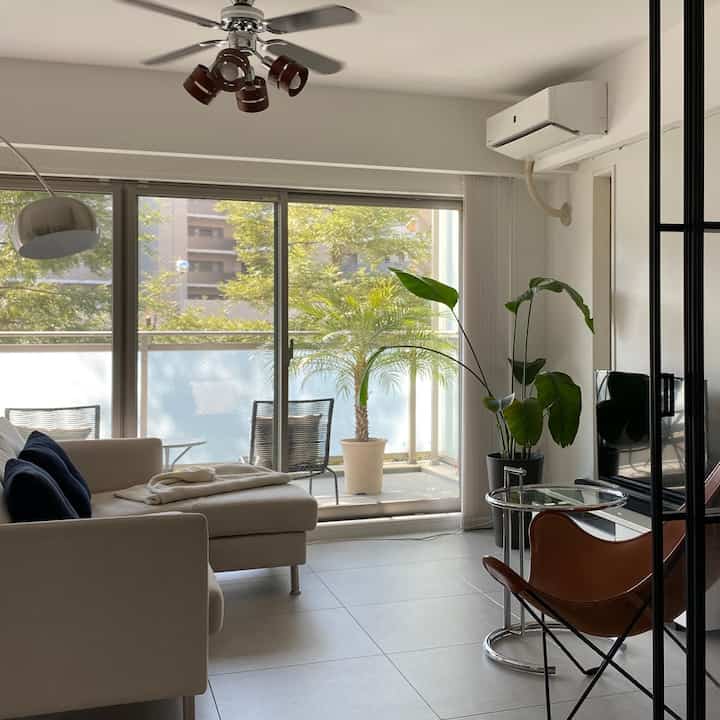 Bright white and beige living room featuring a beige sofa, brown armchair, large plants, and a clean modern interior with ample natural light