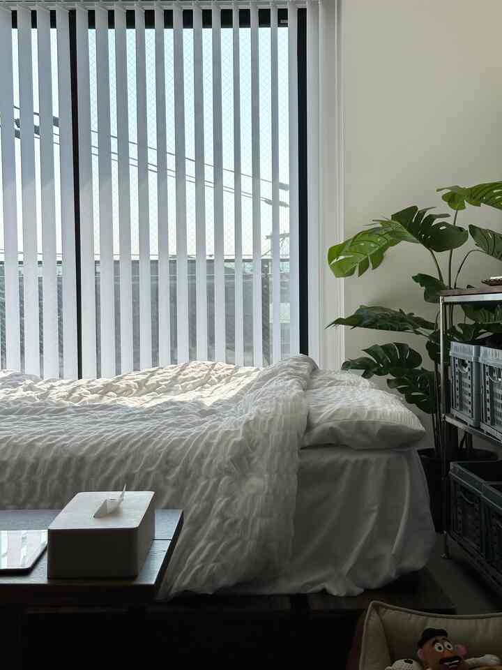 白とナチュラルトーンの寝室、白いベッドカバーとカーテン、植物が配置された清潔感のあるインテリア