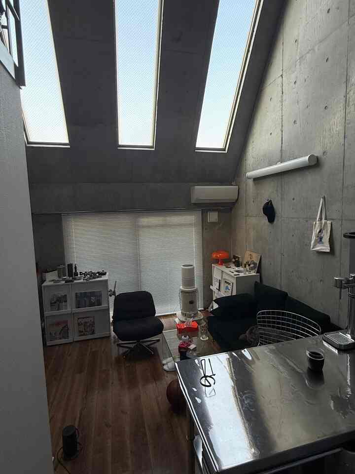 グレーのコンクリート壁と天井、高い天井とスカイライトがあるスペース、黒いソファとナイトテーブルが配置されたモダンな一人暮らしのリビング