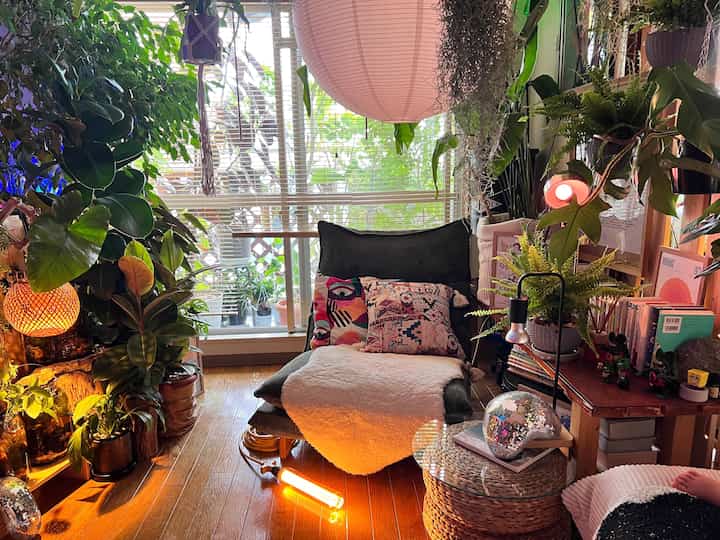 Warm wood tones and abundant green plants fill a long narrow living room featuring a cozy sofa and diverse lighting elements