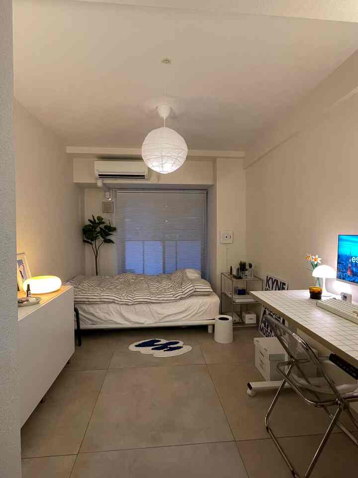 ホワイトとグレーの落ち着いた色調のコンパクトなワンルーム寝室、シングルベッドとデスク、ブラインド、間接照明が配置された雰囲気のあるインテリア