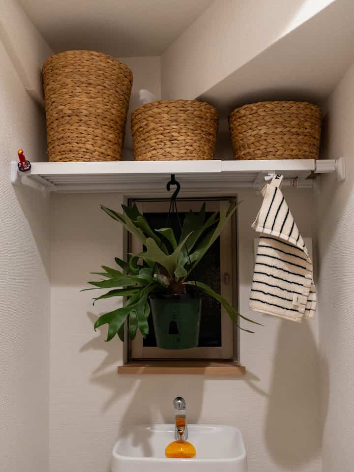 Natural-toned small bathroom with white walls and ceiling, featuring brown wicker baskets on upper shelf and a hanging green plant