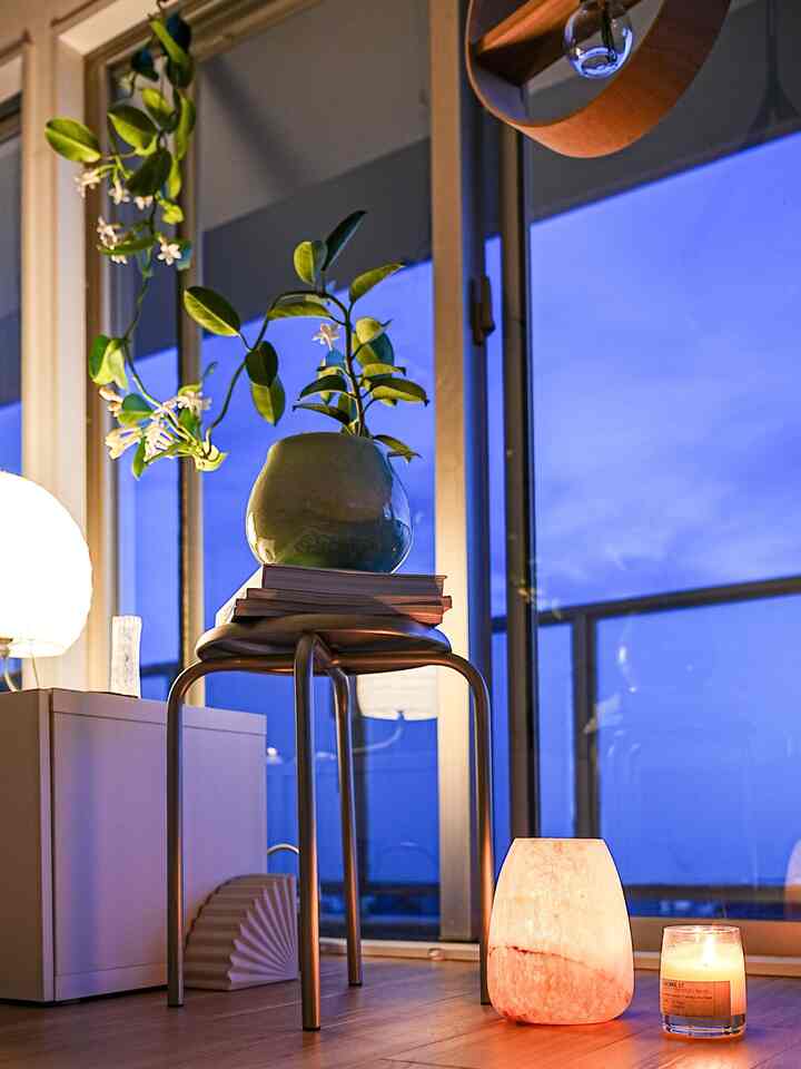 A balcony space with navy blue sky visible through windows, featuring a white shelf and stool, with candles and a plant creating a warm atmosphere