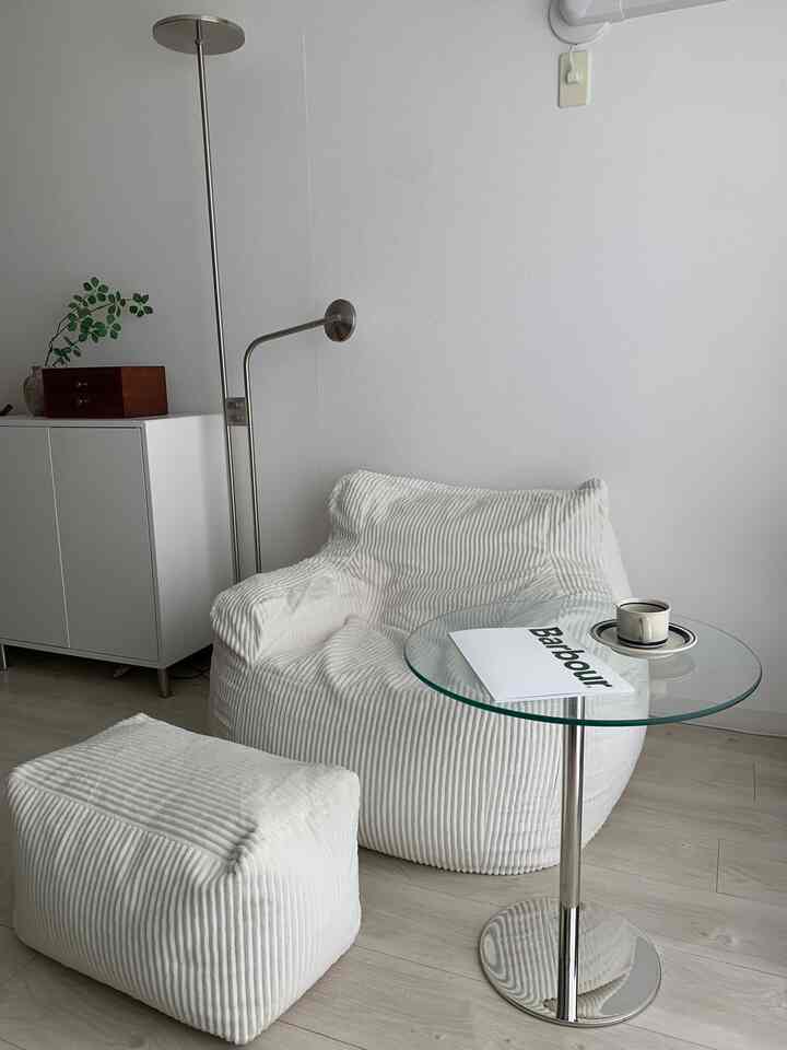 A white and beige toned living room featuring a corduroy bean bag sofa, pouf, glass side table with a minimal and natural atmosphere