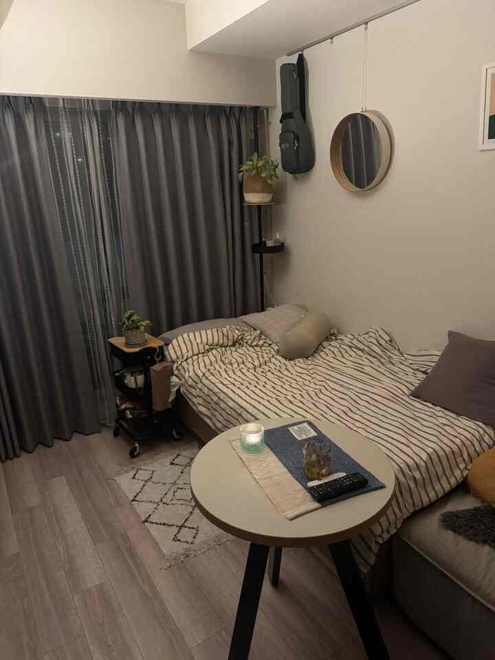 ナチュラルな木目調の床とグレーのカーテンがある一人寝室、ラウンドテーブルと植物が配置されたミニマルで落ち着いた雰囲気のインテリア