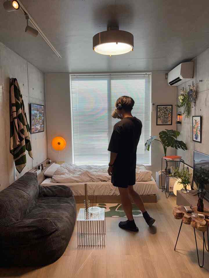 グレーのコンクリート壁とナチュラルな木目調床の1Kワンルーム寝室、ダークグレーのソファと照明、窓際にベッドが配置された落ち着いたインテリア