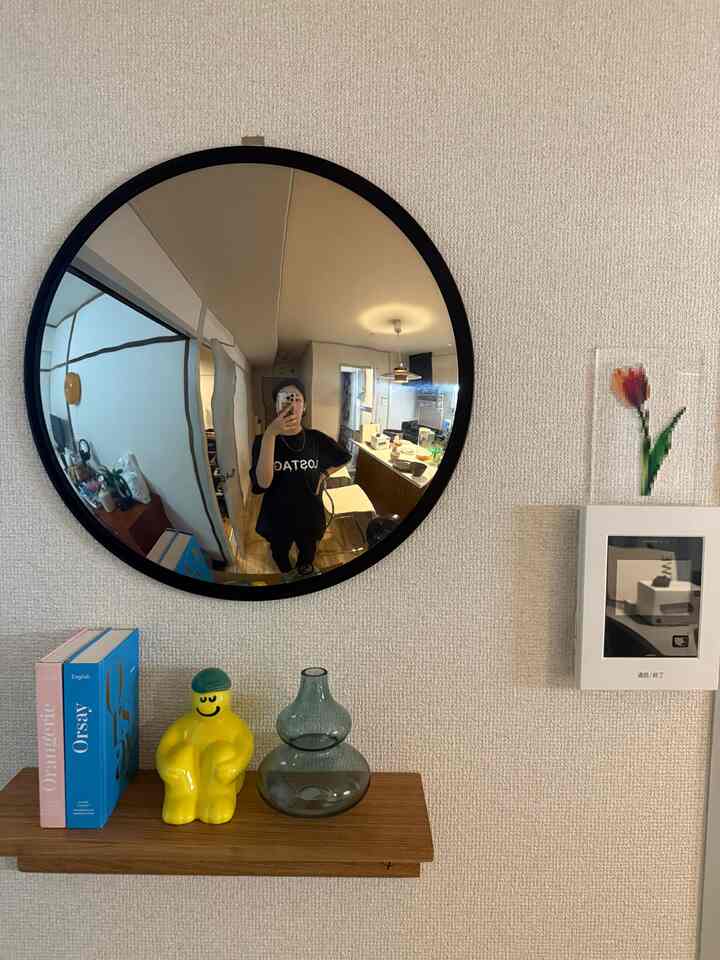 Neutral beige wall featuring a round black-framed mirror and a wood-tone shelf with a yellow ceramic objet and transparent vase