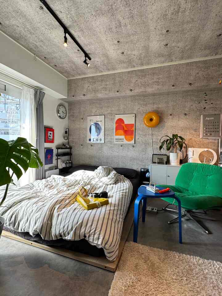 Mid-century modern 1K bedroom with prominent gray concrete wall and ceiling, featuring central bed, green armchair, and blue side table
