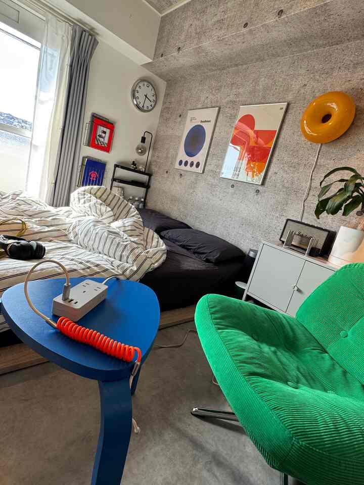 グレーのコンクリート壁と自然光が差し込むミニマルな寝室、グリーンのアームチェアとブルースツールが配置された空間