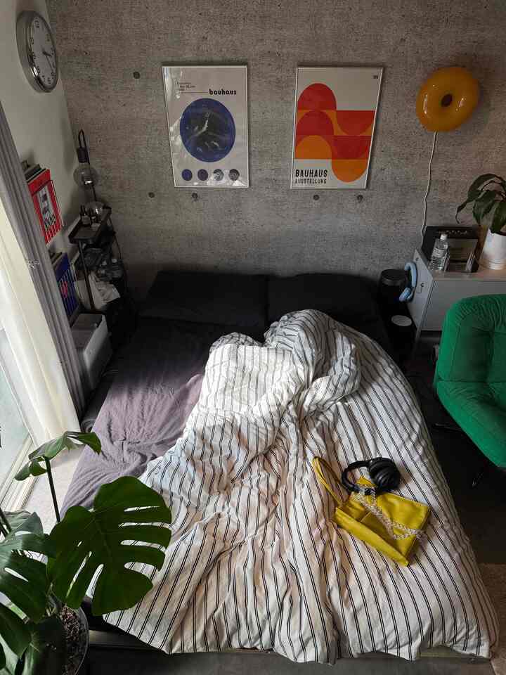 モダンなコンクリート壁と自然光が調和した寝室、中央に大きなベッドと緑のアームチェアが配置された印象的な空間