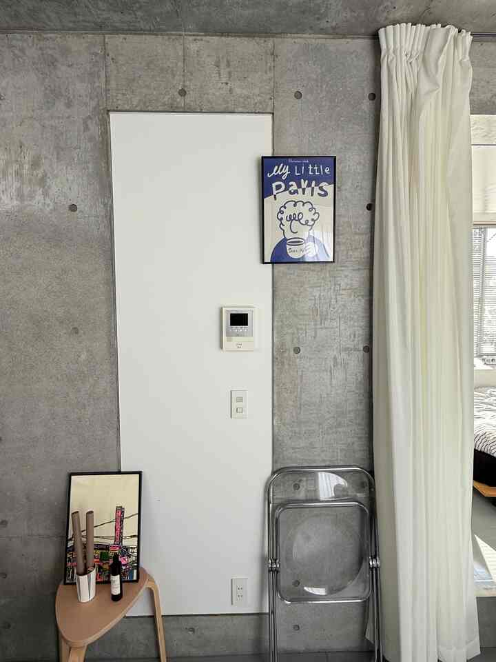 グレーと白のトーンのリビング壁、ポスターと三角形のスツール、折りたたみ椅子が配置されたミニマルな空間