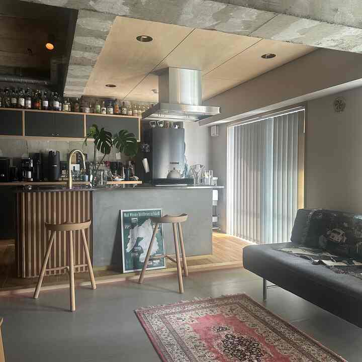 グレーと木目調が調和したモダンなリビングとオープンキッチン、ブラインドとバースツールが配置された雰囲気のある空間