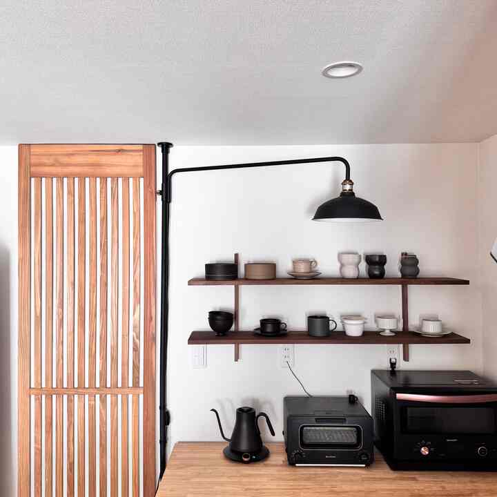 白い壁と木目調のカウンターがあるキッチン、黒い家電と壁の棚に並ぶカップが印象的なホームカフェのある空間