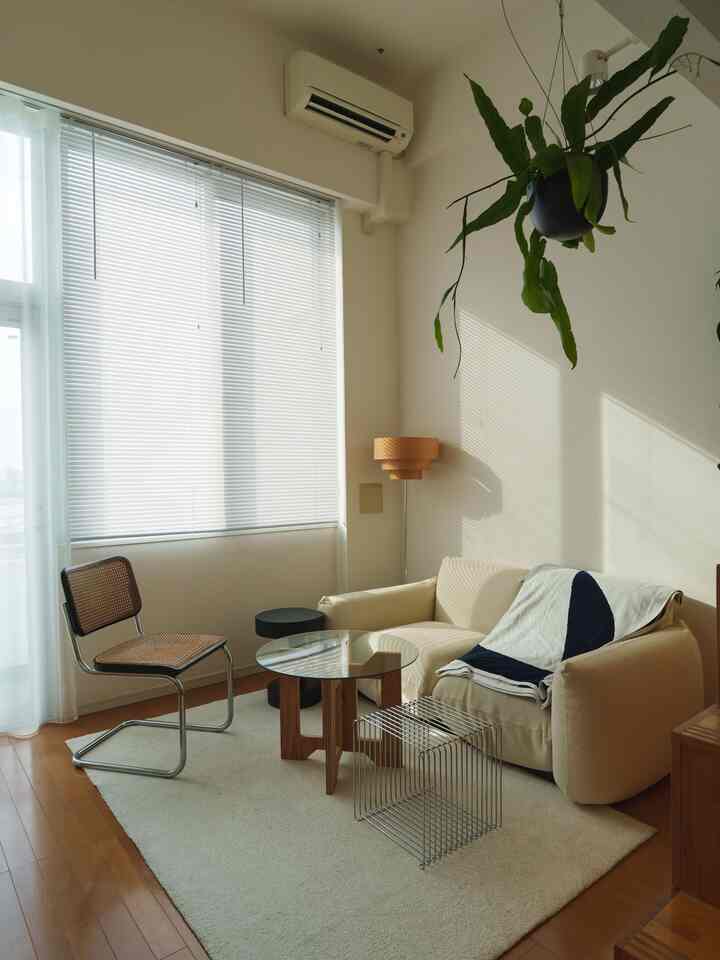 白と木目調の1LDKリビング、クリーム色のソファと特徴的なスツール、ブラインドが配置されたミニマルなインテリア空間