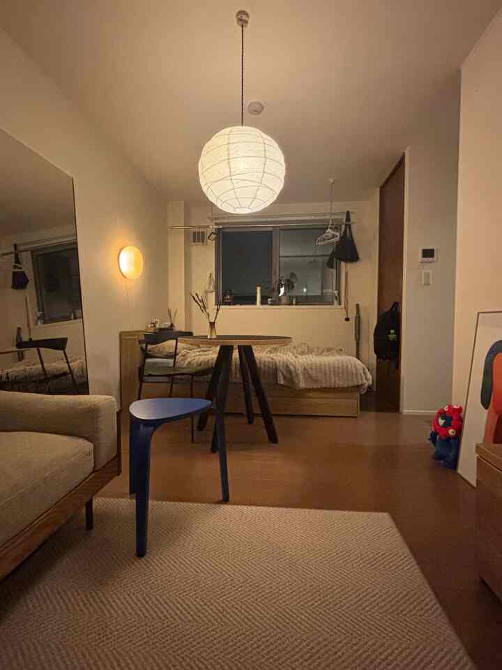 ベージュと木目調のナチュラルな1人暮らし用コンパクトなリビング兼寝室、中央にペンダントライトとブルースツールが配置された印象的な空間