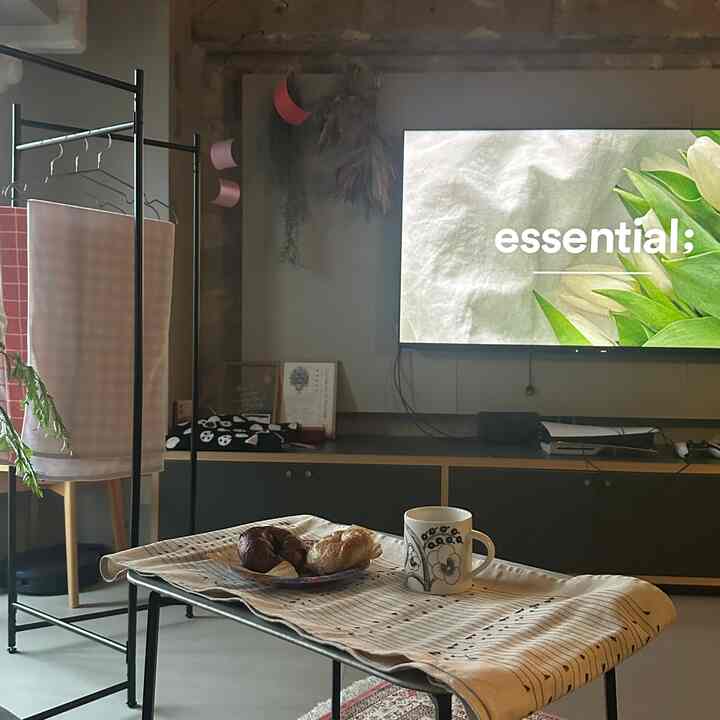 ベージュと黒の色調のリビング、コートラックやテレビ台が配置され、テーブルクロスのかかったテーブルの上にベーグルとマグカップが置かれた雰囲気のあるおうちカフェ空間