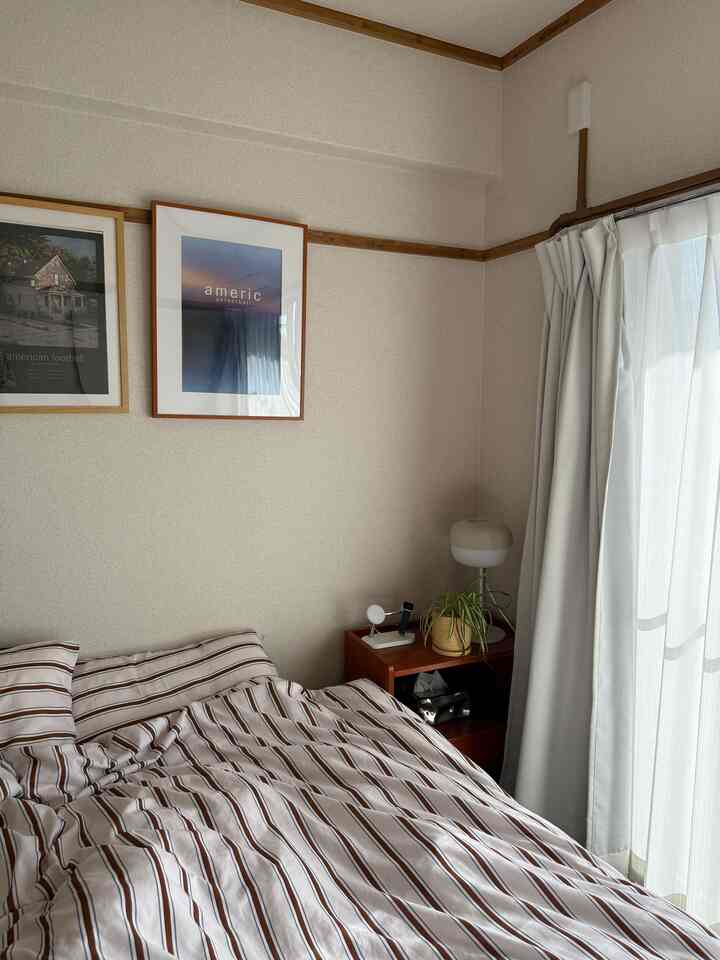 白い壁とカーテン、ストライプの寝具がある6畳の寝室、ナイトテーブルに植物が置かれた落ち着いた空間