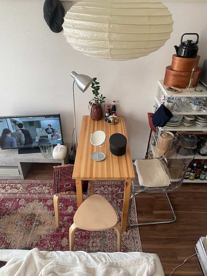 ナチュラルな色調の6畳の部屋、木製のデスクとスツール、透明な椅子、ペンダントライトが配置されたシンプルなホームオフィスの空間