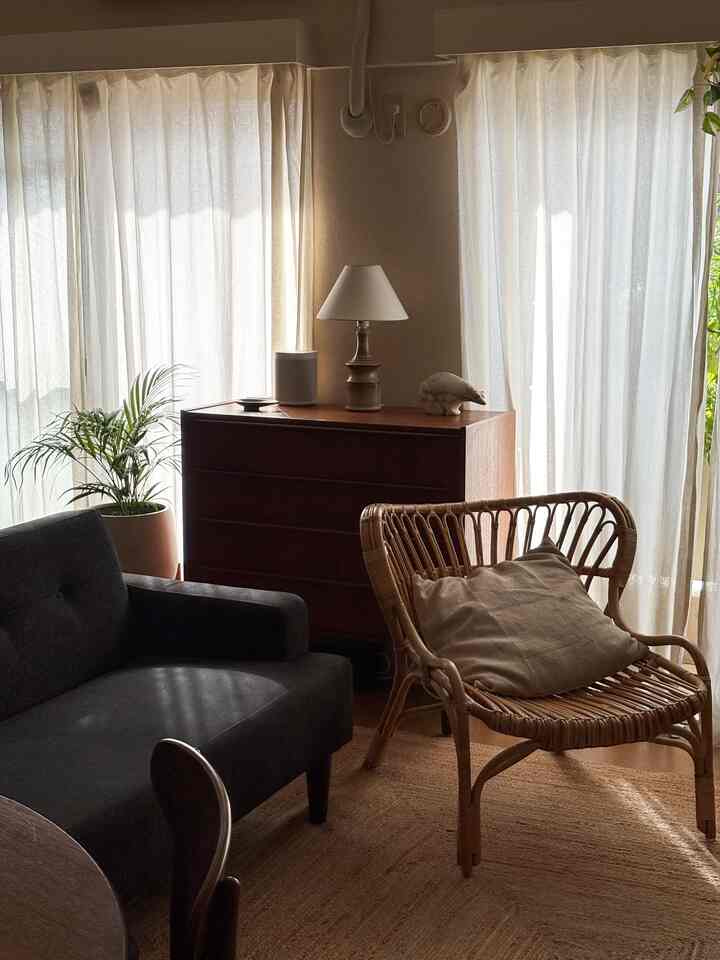 ベージュとブラウンの色調のリビング、右側にラタンチェア、左側に黒いソファ、中央にチェストとテーブルランプが配された雰囲気のある空間